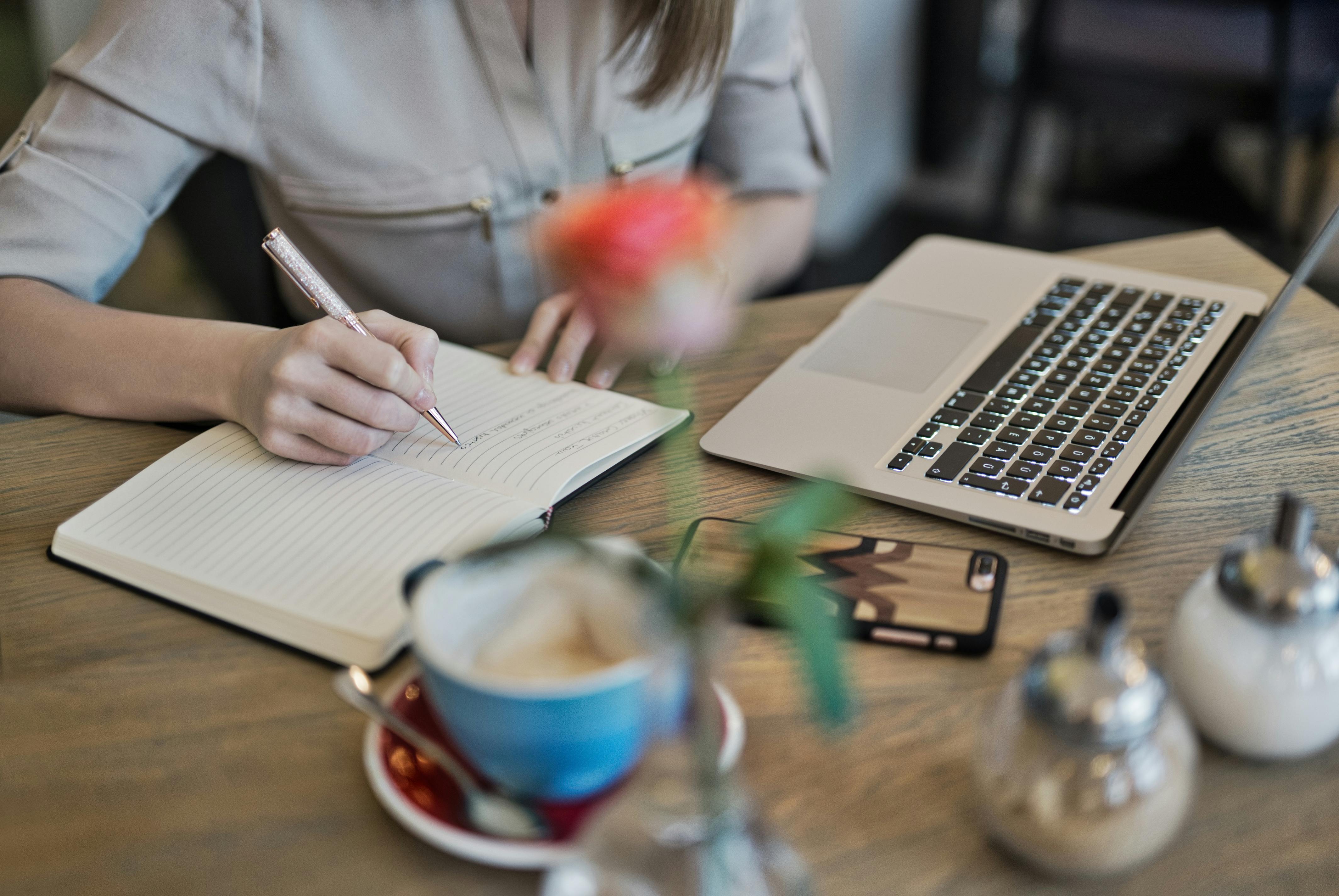 Mujer joven escribiendo en su cuaderno, junto a su ordenador portátil, sus gafas y una taza de café