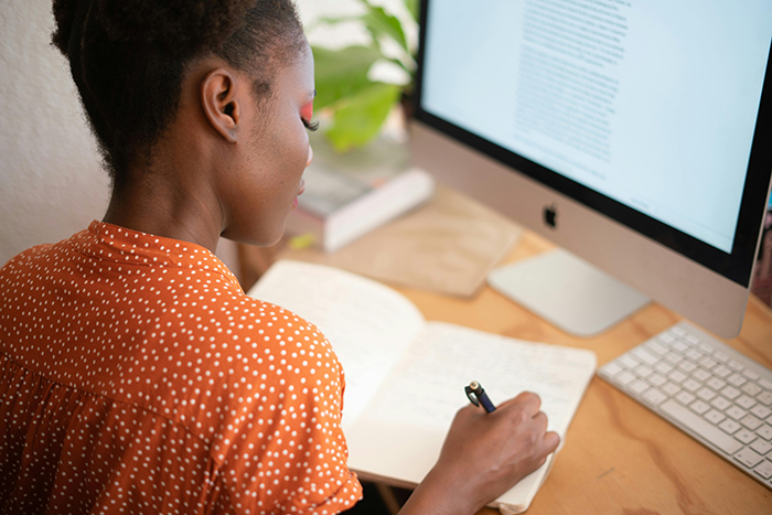 Persona escribiendo en un cuaderno con una computadora delante